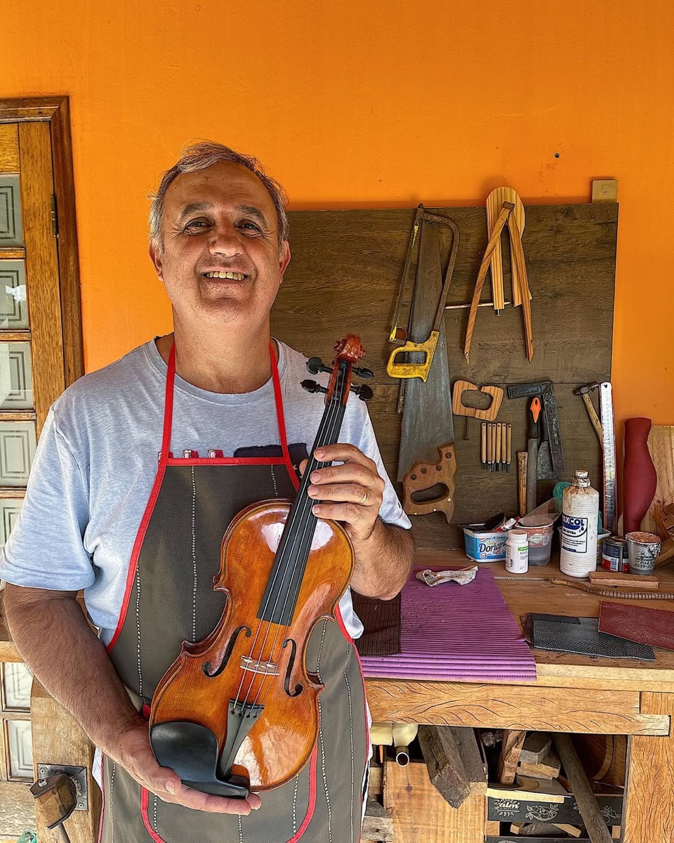Marcos Belvis, luthier, na oficina com avental e um violino nas mãos, em frente à bancada e ferramentas.